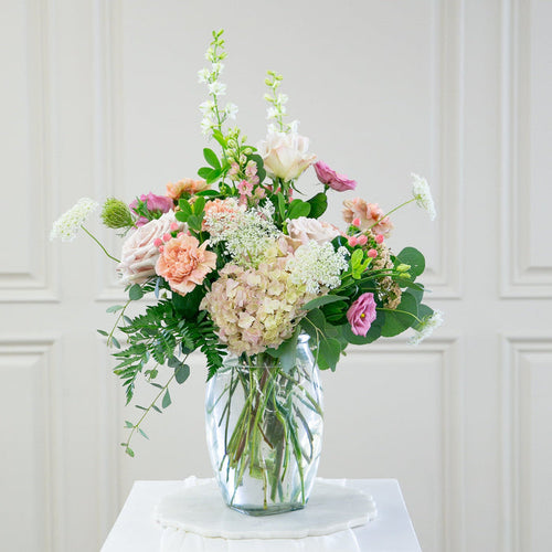 Bouquet of flowers in a clear vase on a white pedestal against a white paneled wall.