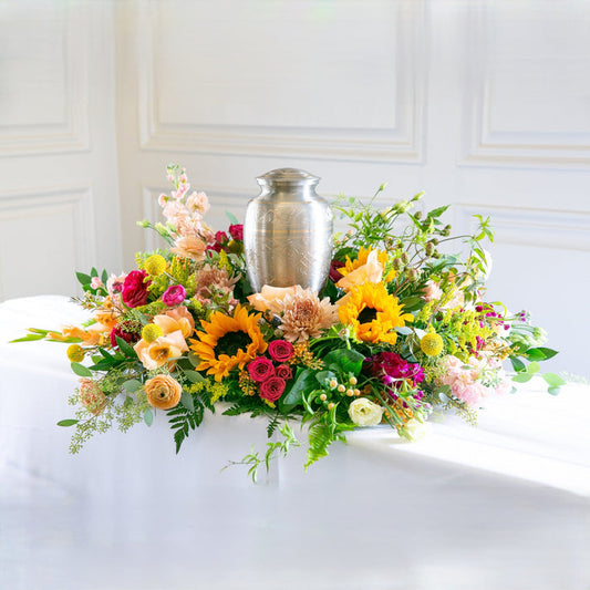 Silver urn surrounded by colorful flowers on a white surface with a white paneled wall background